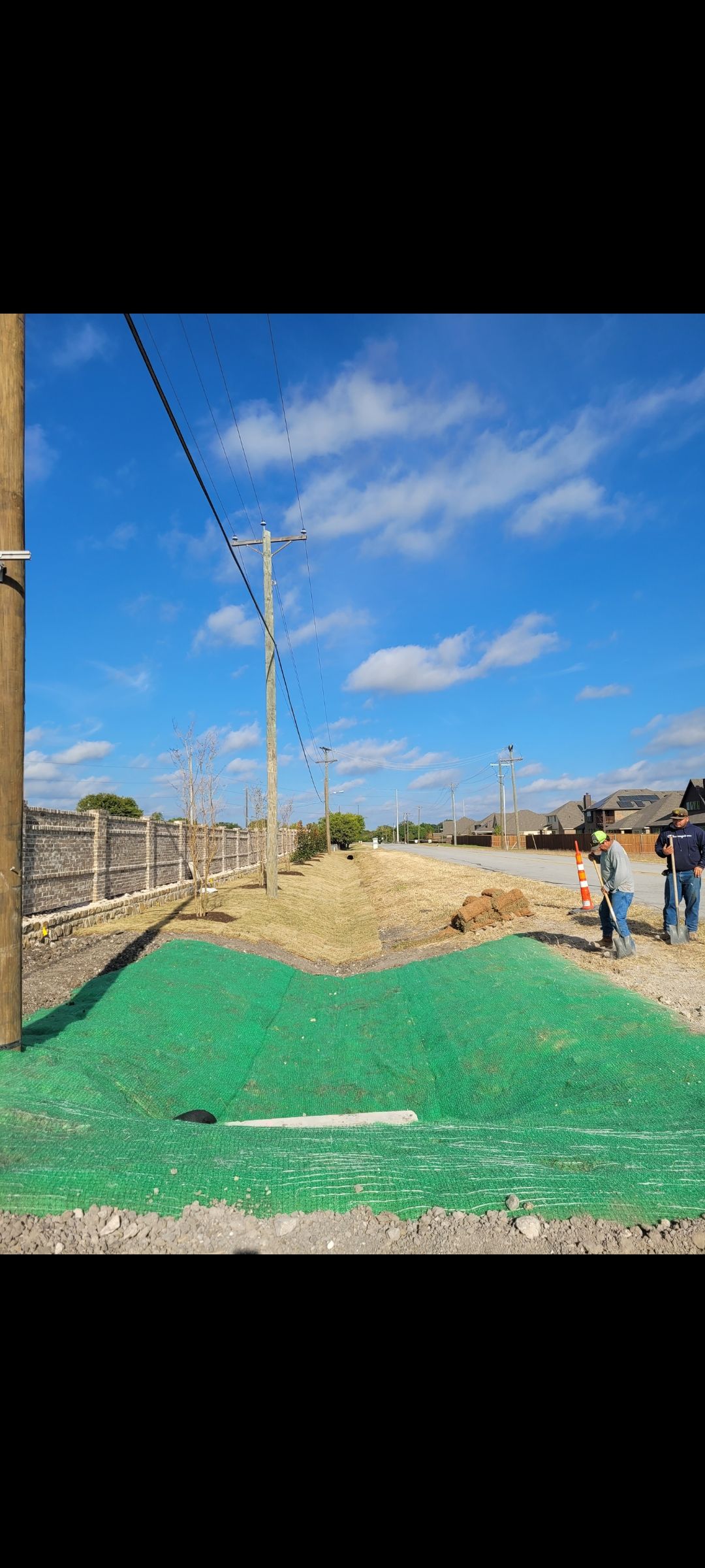 Outdoor construction site with green tarp, dirt mound, power pole, and workers under a blue sky