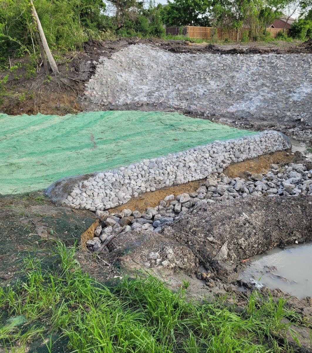 Green tarp-covered embankment beside a gravel pile and muddy water in a rural construction site