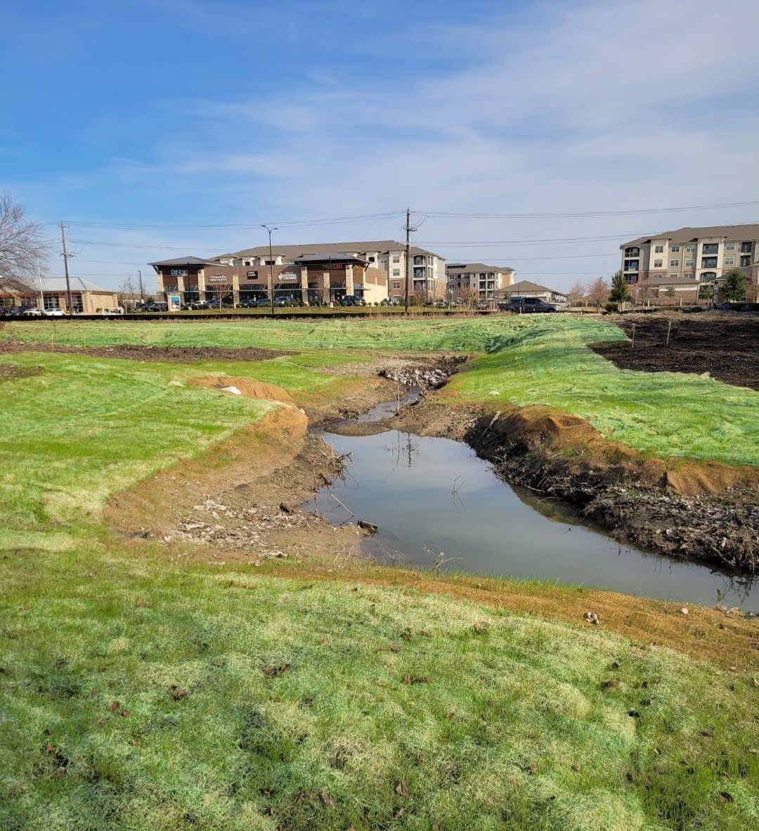 Grassy field with a narrow stream, under blue sky, with apartment buildings in the background