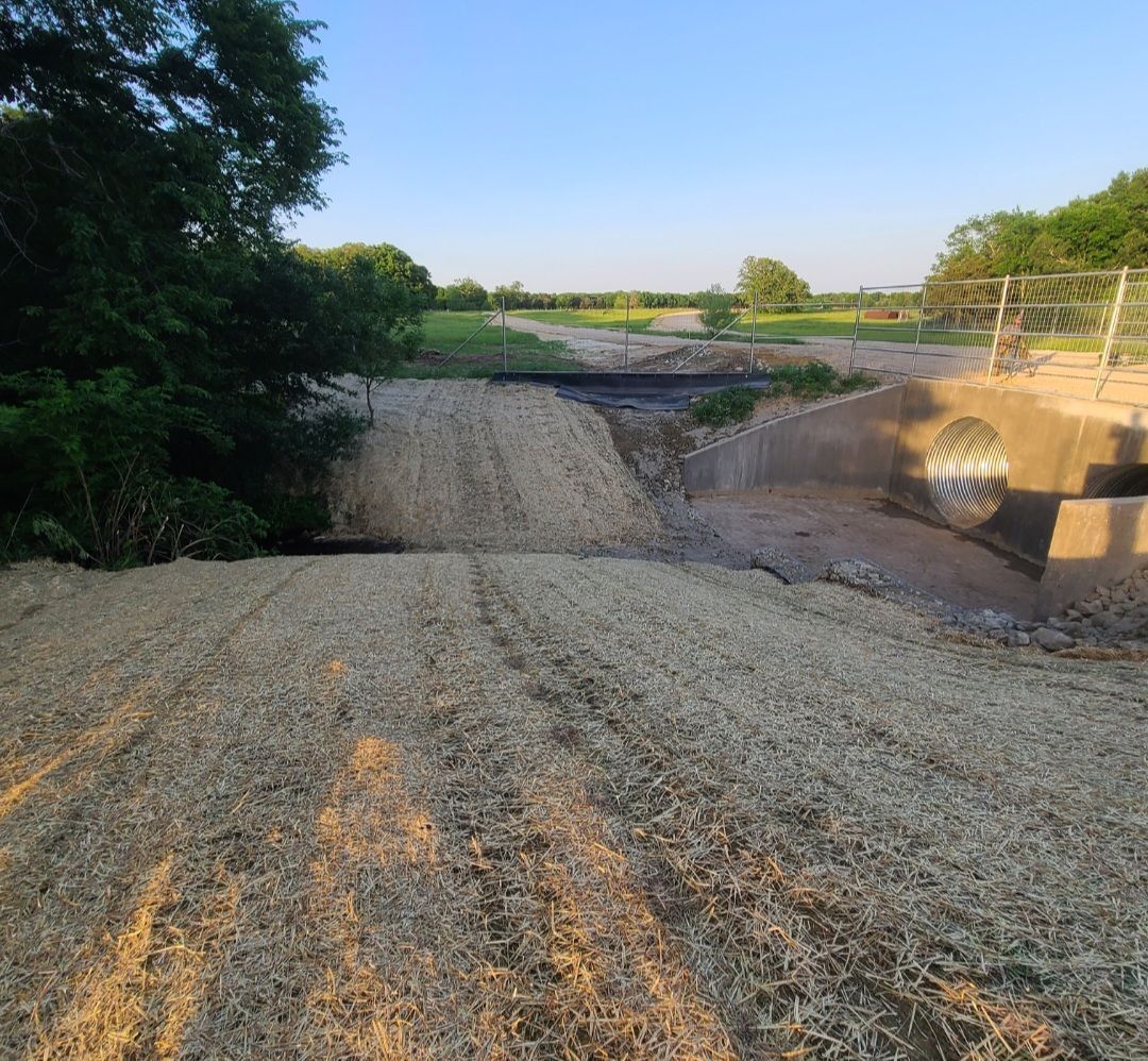 Gravel road washed out by erosion under a small bridge, with trees and fields in the background