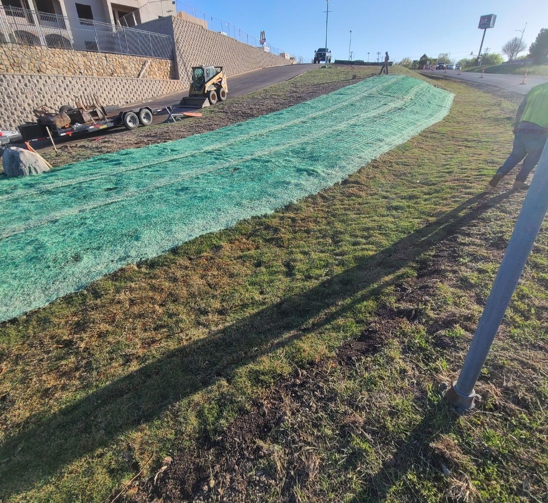 Green netting covers a grassy slope beside a road, with a stone wall and equipment on the left.