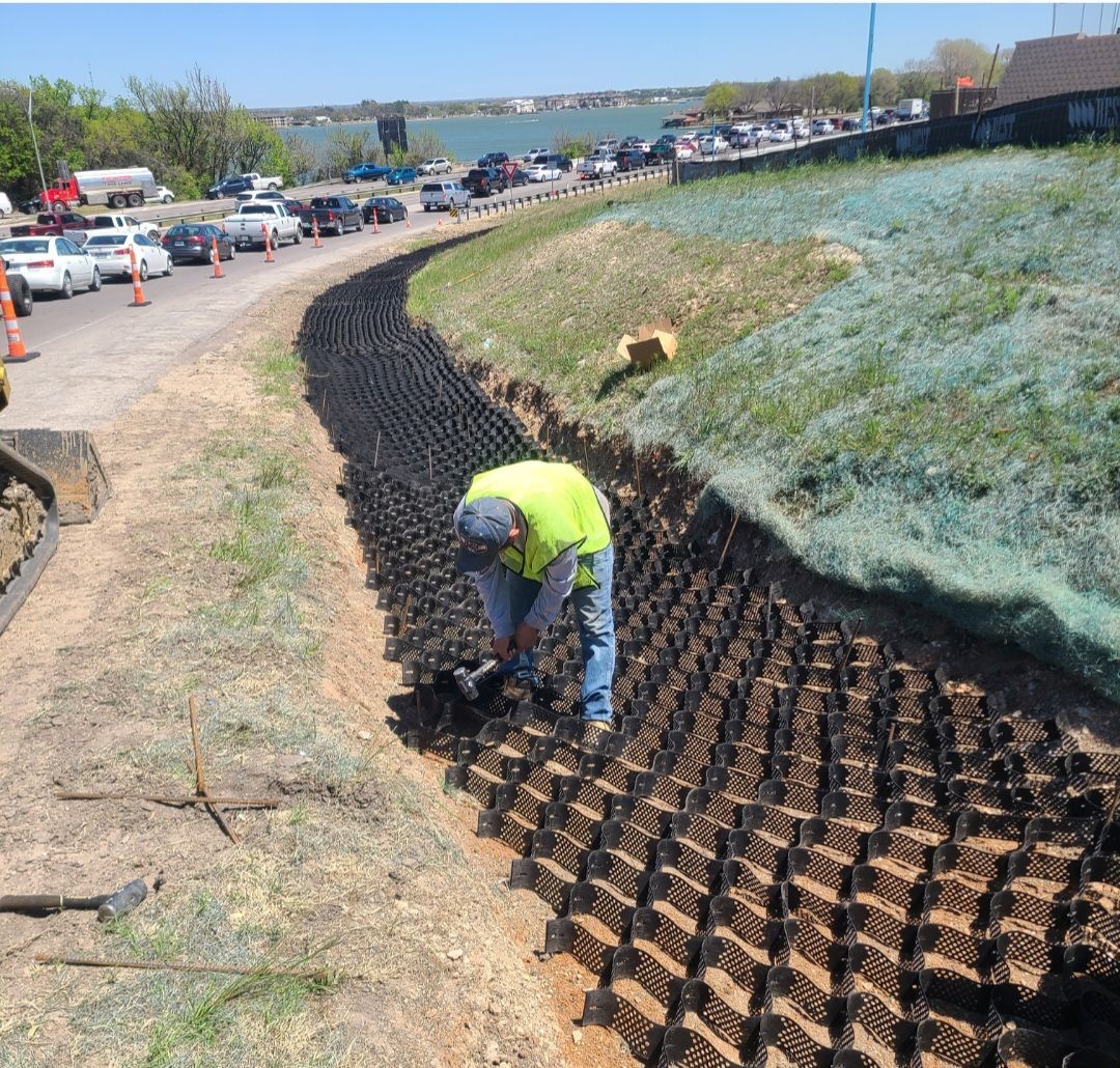Worker installing erosion-control matting on a sloped roadside embankment beside a lake.