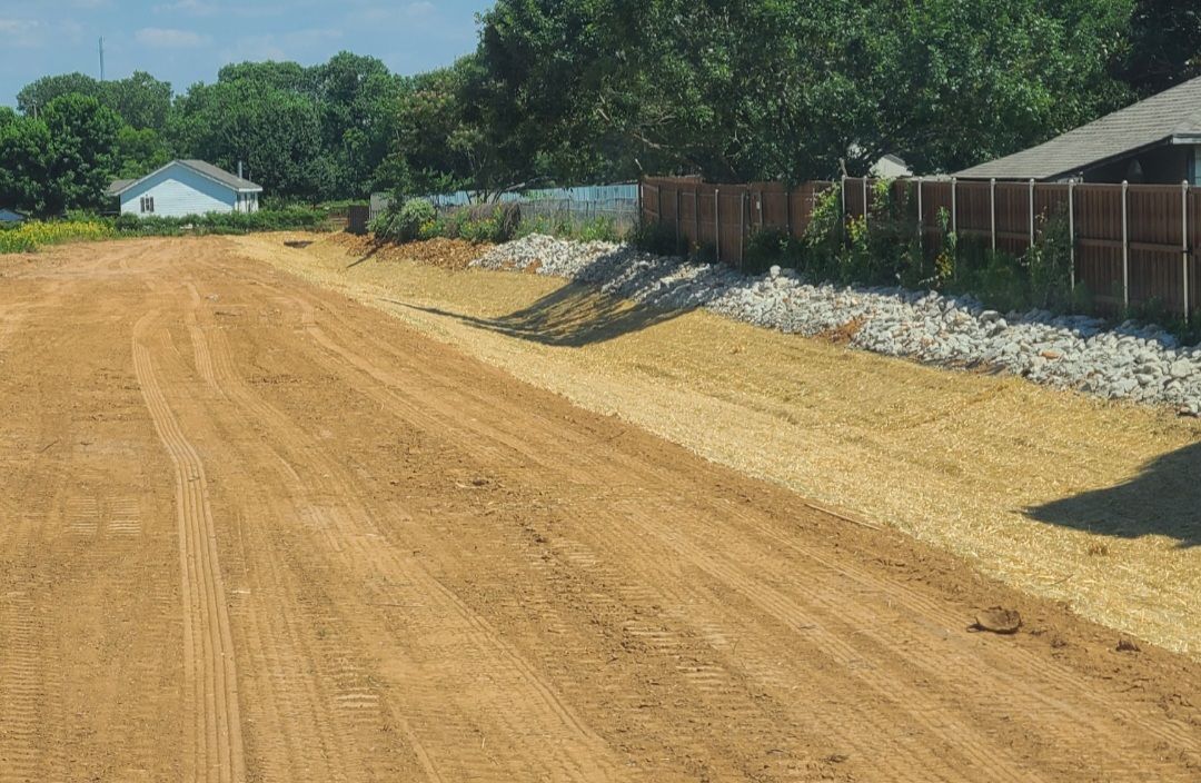 Gravel road under construction beside a fence, with houses and trees in the background.