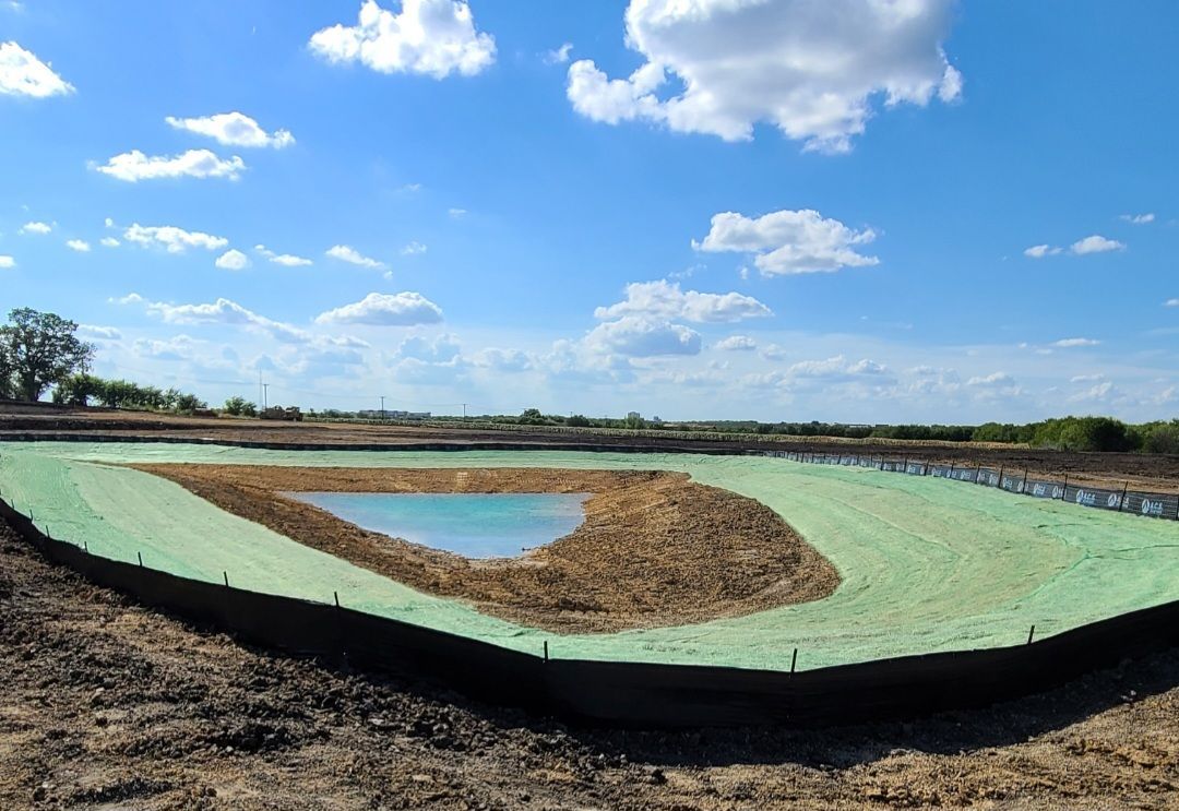 Round earthen pond with green lining and a small blue water pool under a bright cloudy sky.