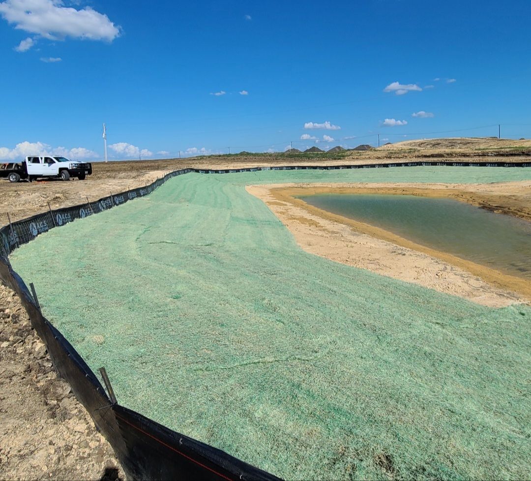New lined pond with green water, sandy embankment, and trucks in the background under a blue sky