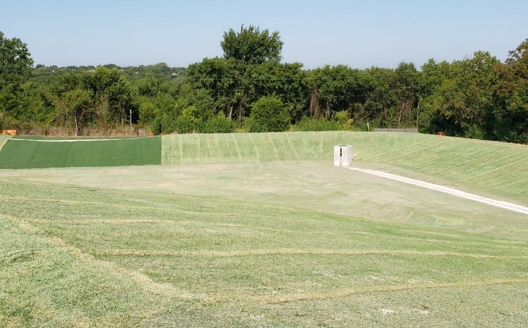 Grassy sports field with a small white structure, green fencing, and trees in the background