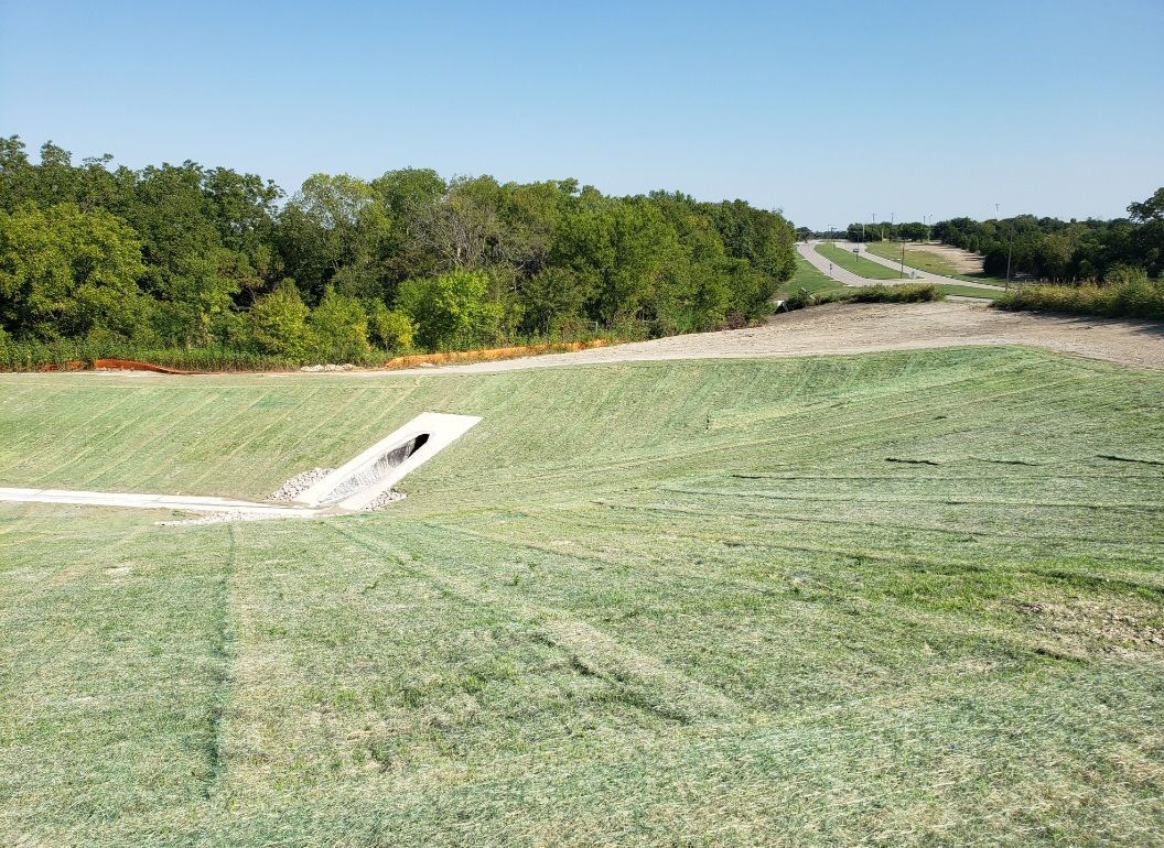 Grassy hillside with a concrete drainage channel beside a tree line under a clear blue sky