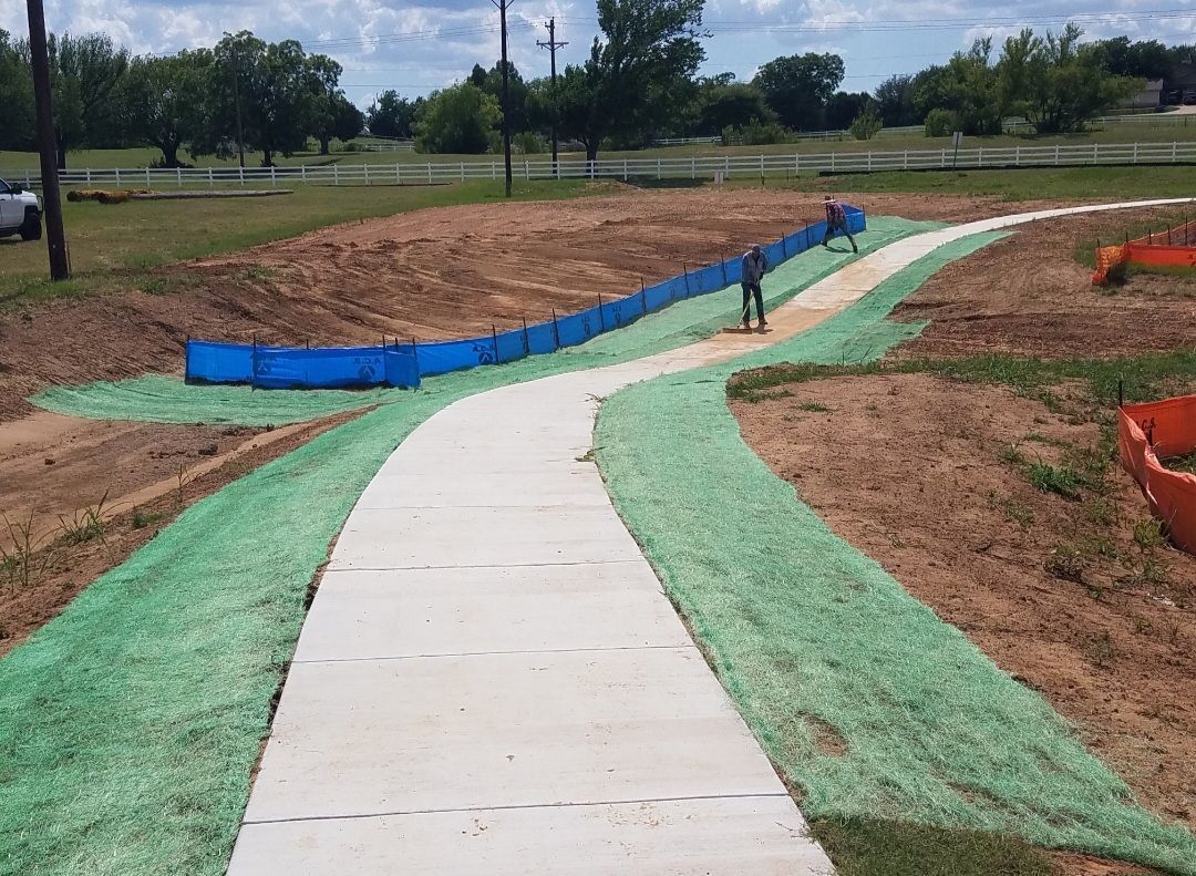 Curving concrete walkway under construction, bordered by green erosion matting and red dirt
