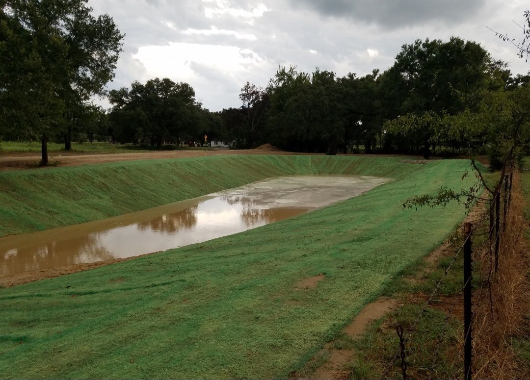 A muddy drainage ditch runs through a grassy field under a cloudy sky.