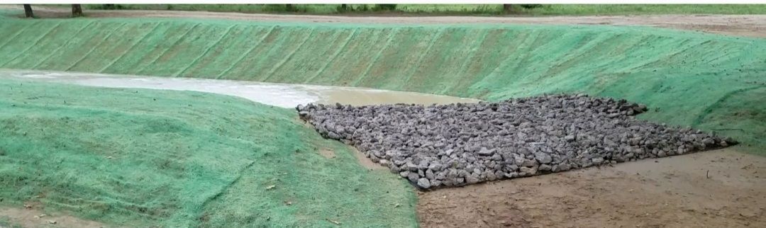 A green-lined irrigation channel with a pile of gray rocks forming a bank or embankment.