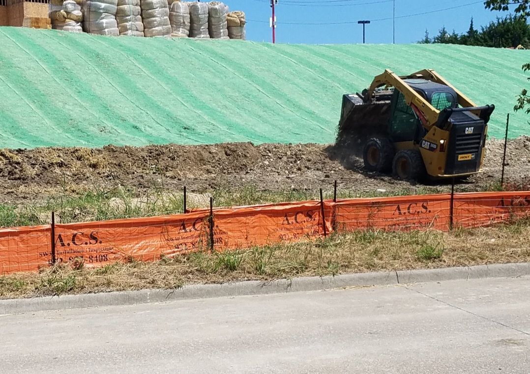 Small excavator beside a green-covered slope and orange safety fence along a roadside