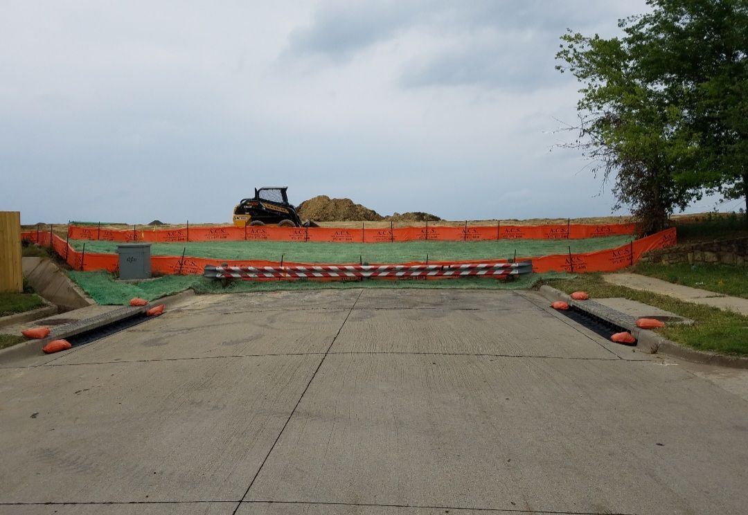 Road blocked by orange barriers across a concrete path near a grassy field and trees.