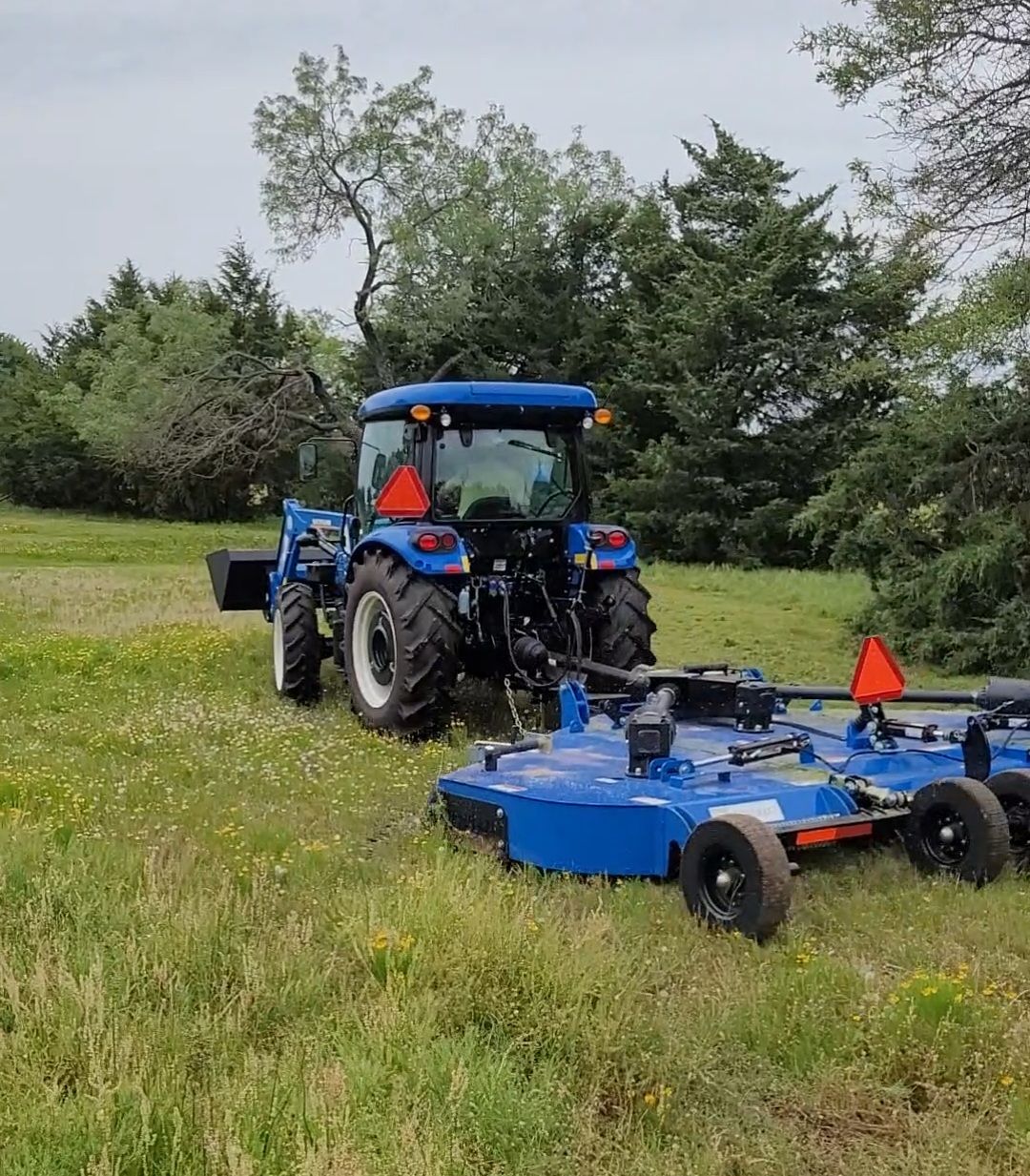 Blue tractor pulling a blue mower on a grassy field, with trees in the background.