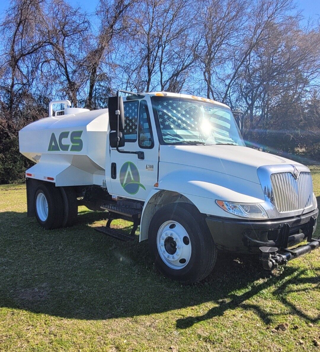 White ACS water tanker truck parked on grass in a sunny field with trees in the background