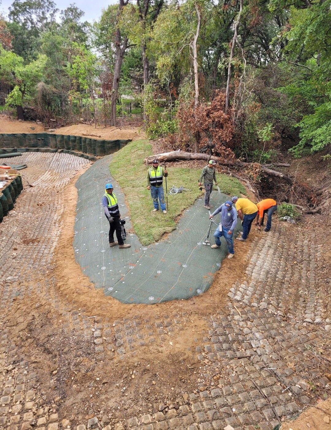 Workers paving a curved path on a hillside, with green mesh and stone blocks in a wooded area.