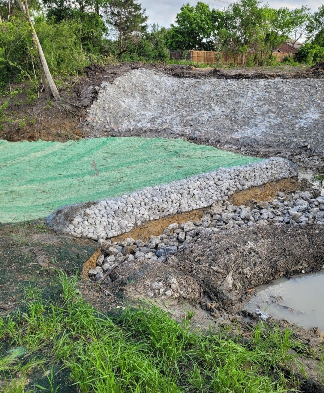 Gravel embankment with green mesh cover beside muddy water and grassy field