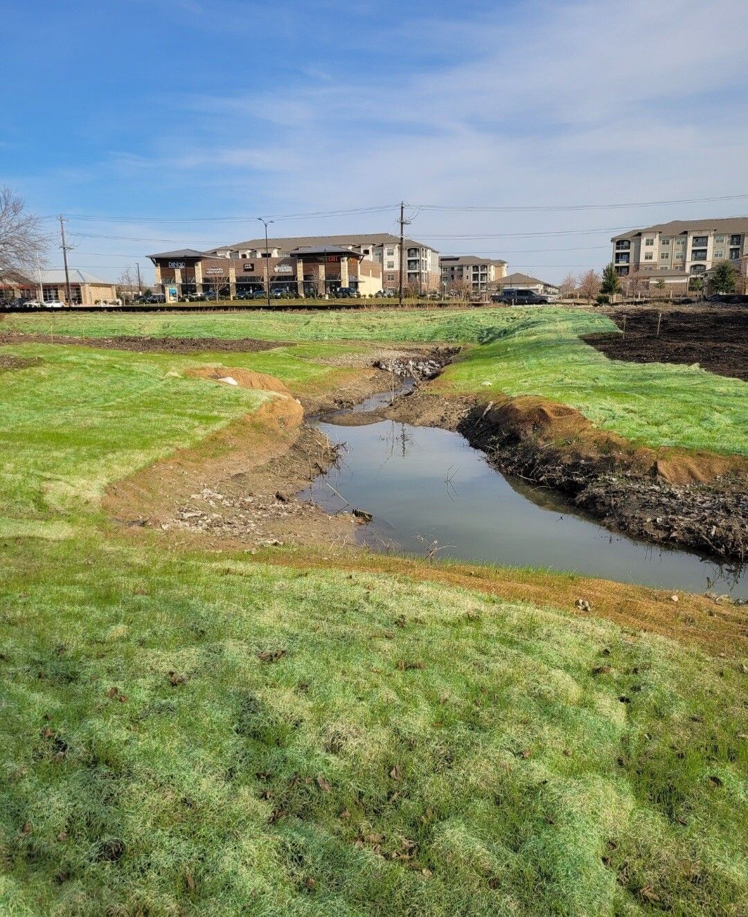 Grassy field with a narrow stream, under construction near apartment buildings on a clear day