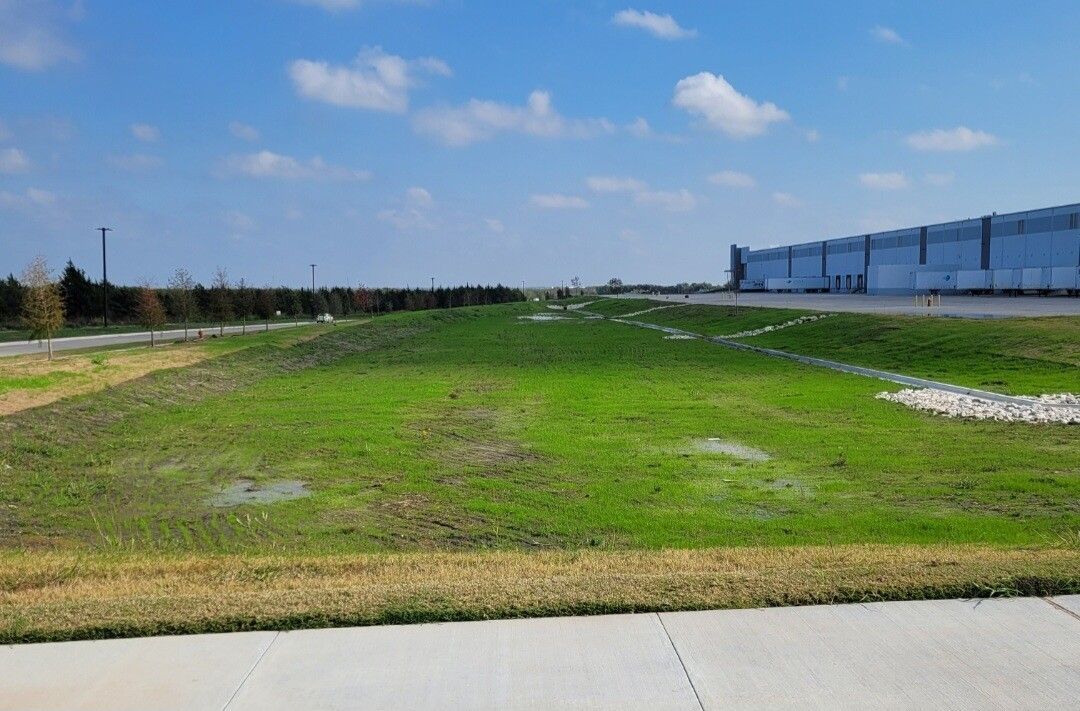 Grassy vacant lot beside a concrete sidewalk, with industrial buildings under a blue sky.