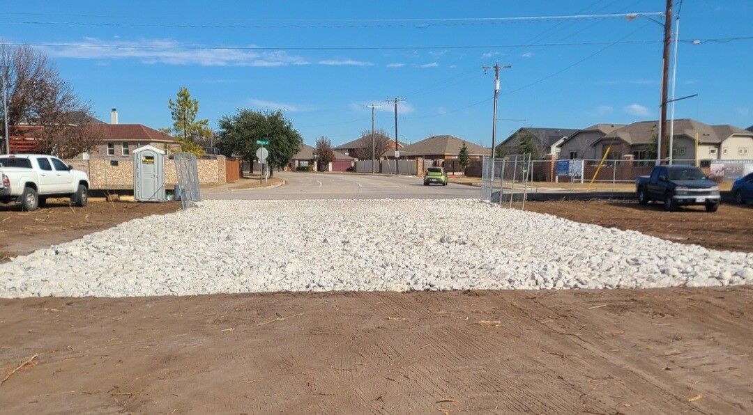 Gravel road base in a residential street under construction, with houses and parked vehicles on both sides.