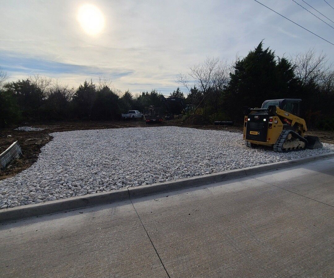 Construction site with a gravel bed and a yellow skid steer beside a road under a cloudy sky