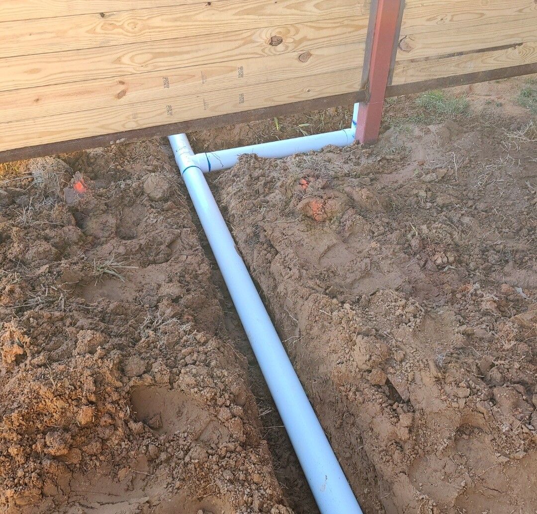 White PVC pipe connected to a red vertical pipe beside a wooden wall in a dirt trench