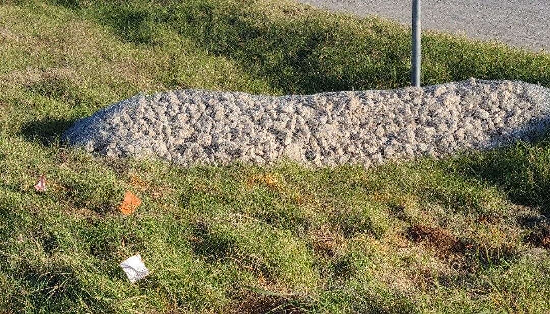 Grassy roadside embankment with a long gray rock pile and a metal post.
