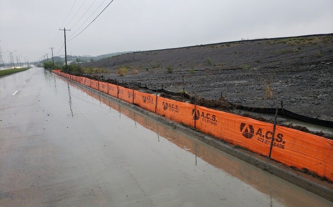 Orange silt fence along a wet roadside beside a dark gravel embankment under overcast skies