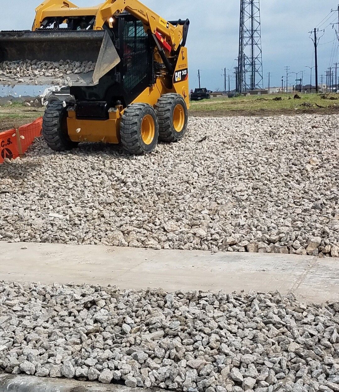 Yellow skid-steer loader on gravel near an industrial site with power lines.