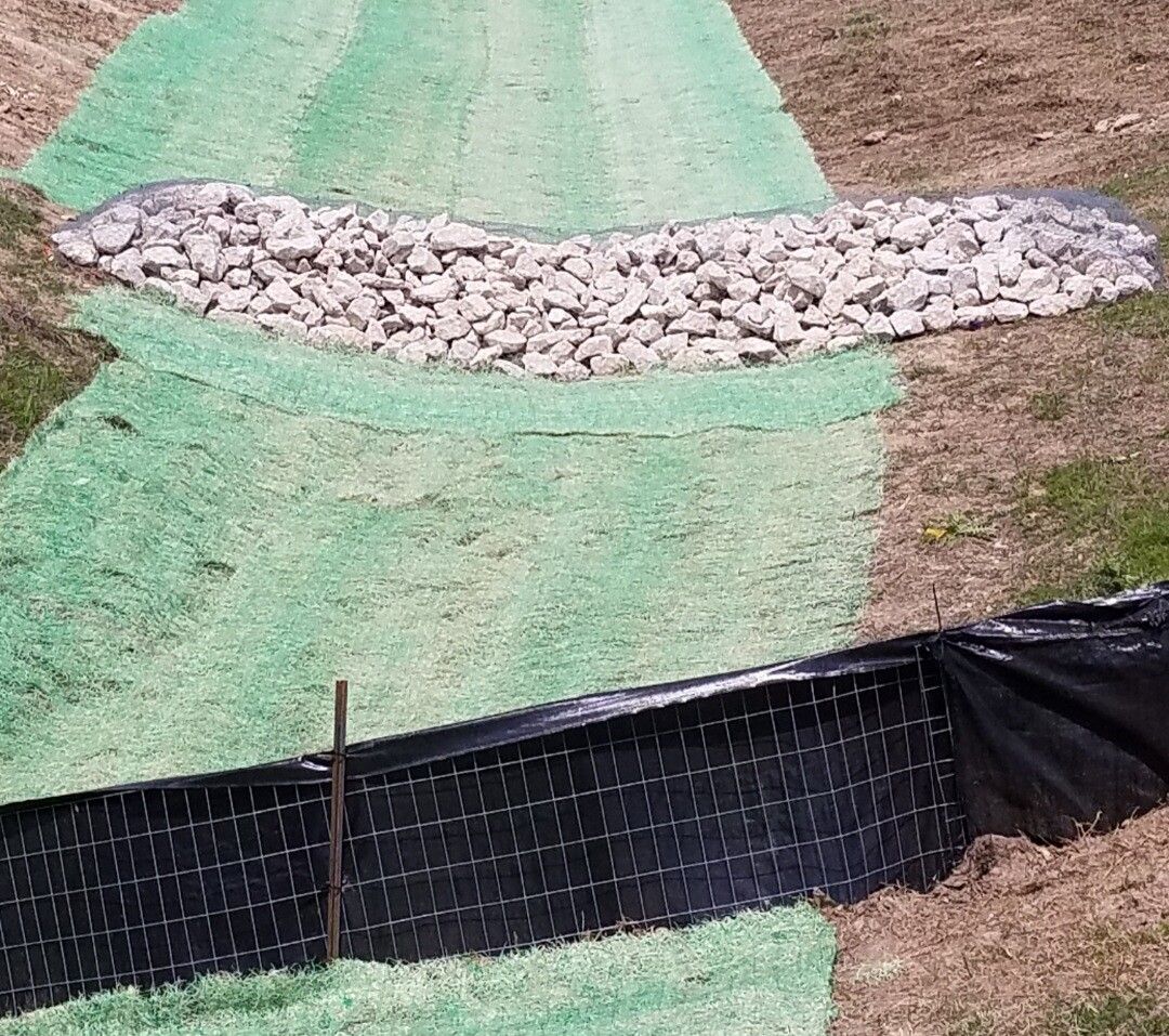 Green erosion-control fabric and black silt fence on a dirt slope with a white rock-lined drainage swale.