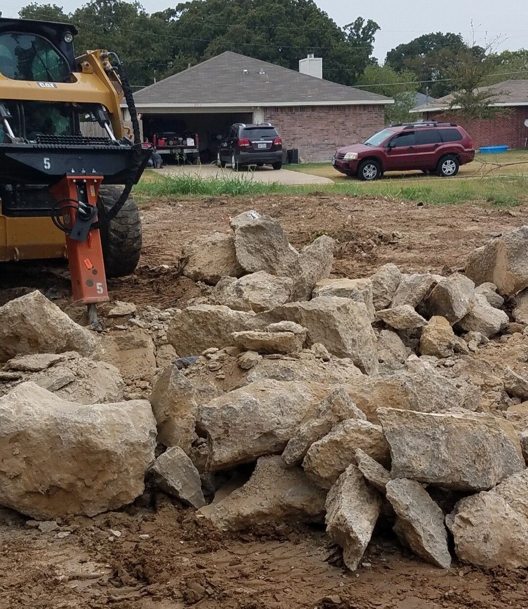 Excavator beside a pile of large rocks at a construction site with houses and parked cars in the background