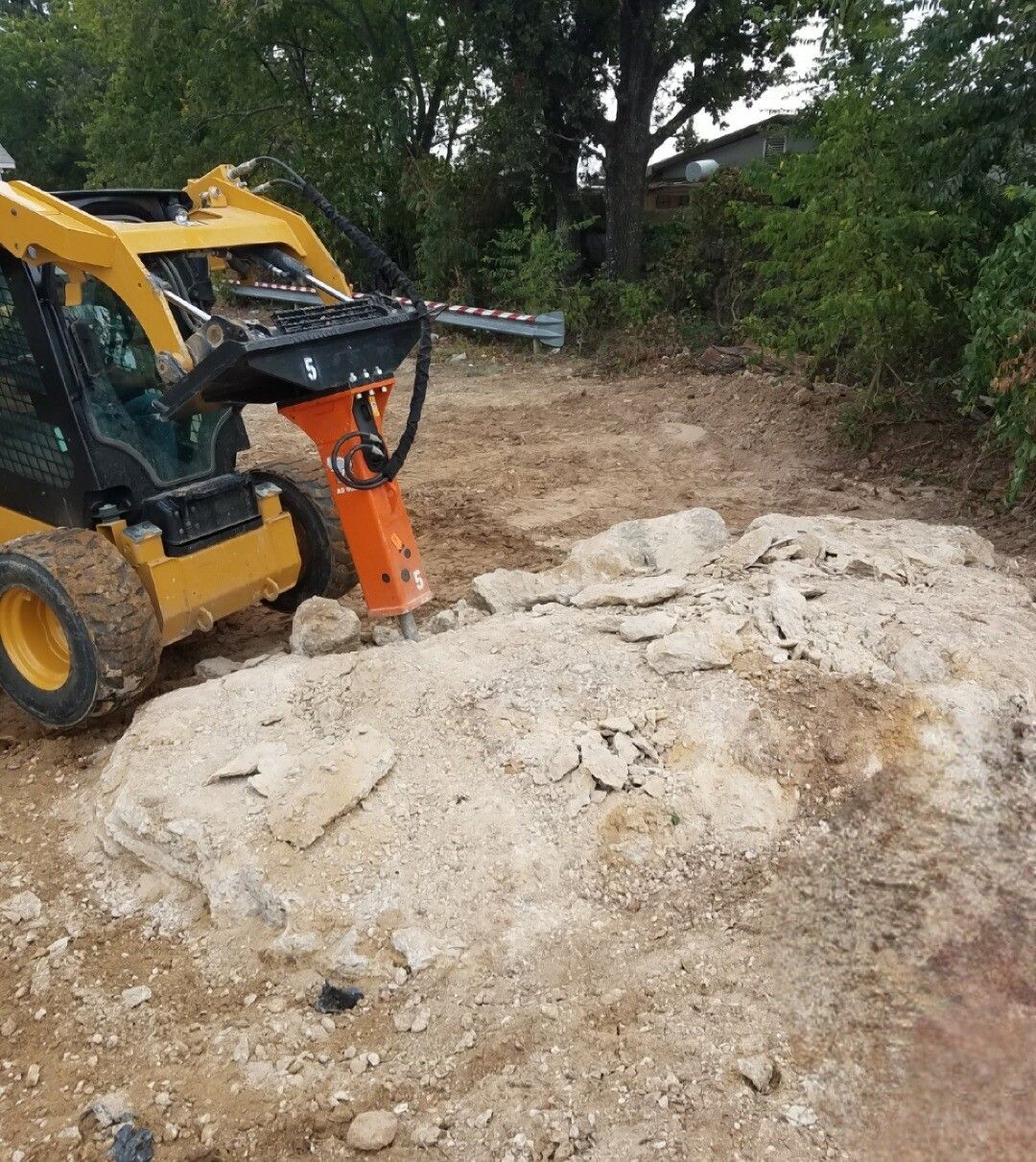 Yellow skid steer with orange attachment clearing dirt on a rocky construction site.
