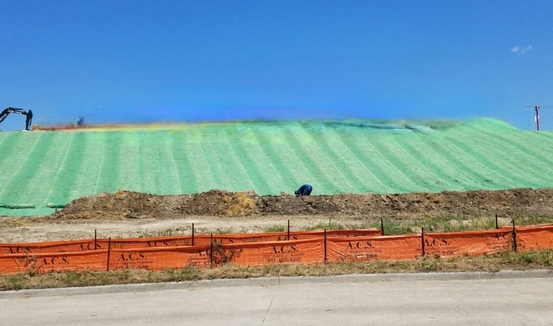Construction site with a green-covered slope, orange barriers, and blue sky above a road