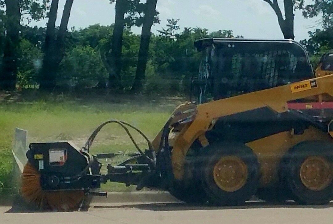 Yellow skid steer towing a black attachment along a roadside with trees in the background