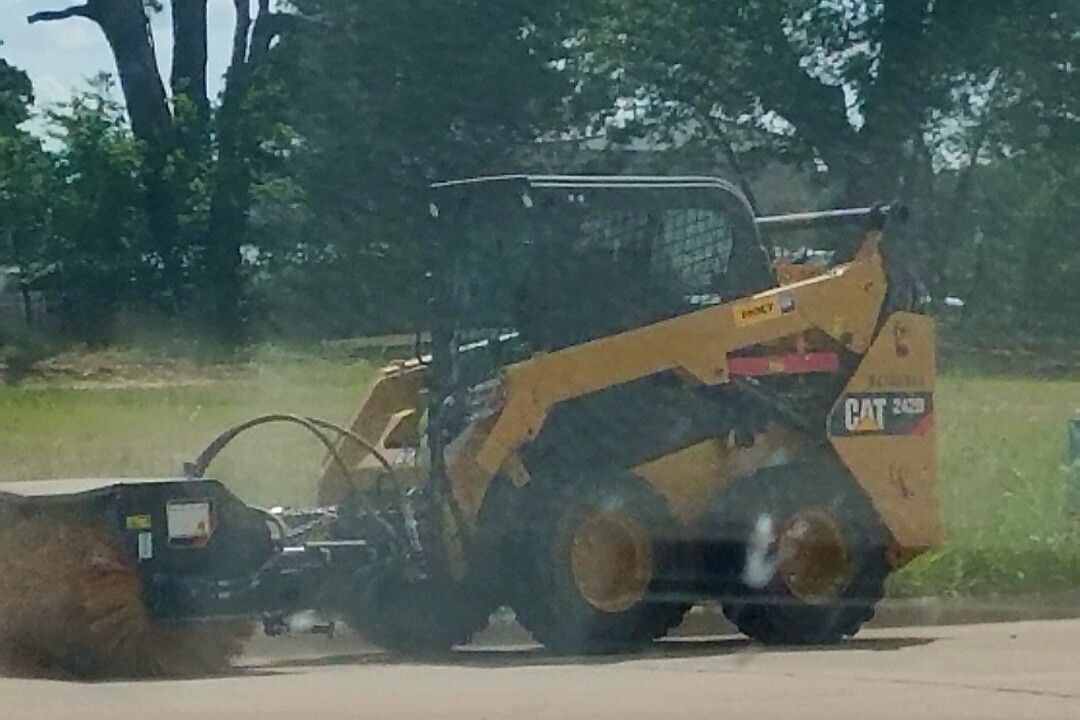 Yellow skid steer with front attachment working on a roadside beside a grassy field