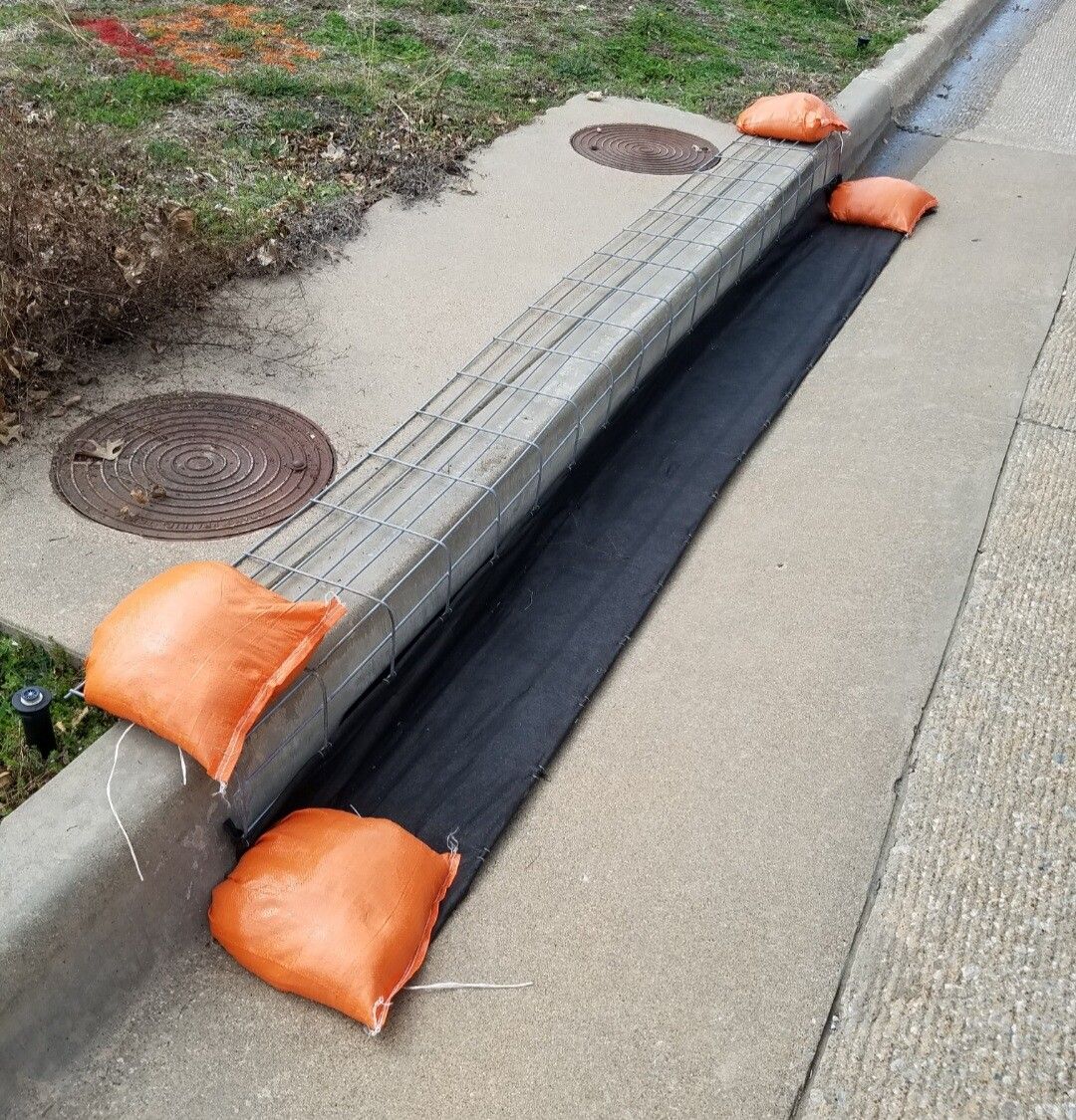 Flooded curb drain covered with black silt barrier and weighted by orange sandbags on a street edge