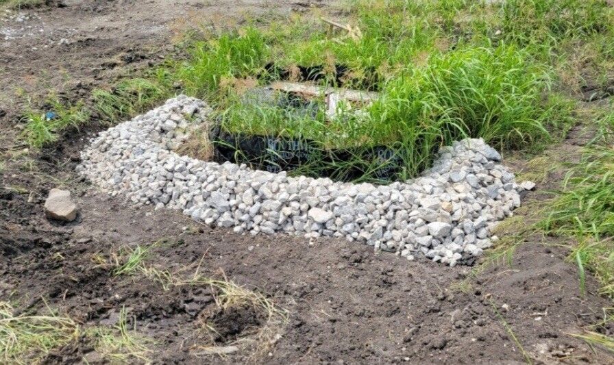 Small circular pond or pit lined with white rocks, surrounded by grass and bare dirt.