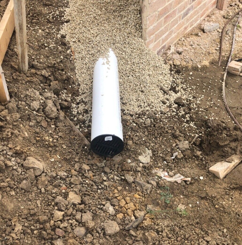 White pipe lying in a dirt trench beside a brick wall and gravel.