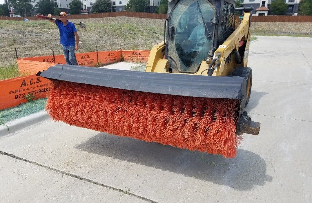 A skid steer with a red sweeping brush cleans a concrete surface near a fenced area.