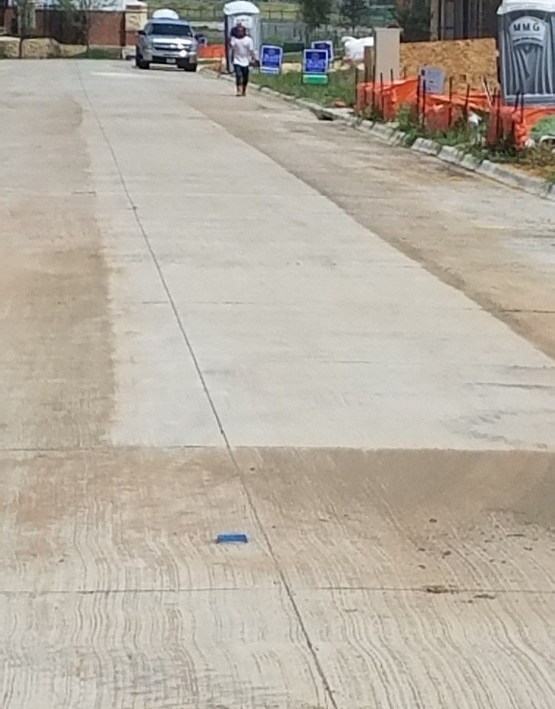 Concrete road under construction with orange fencing, a parked car, and a person walking in the distance.