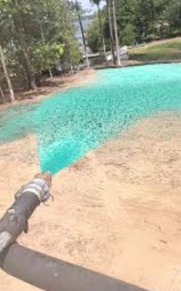 Large hose spraying turquoise water across a sandy area near trees and a road