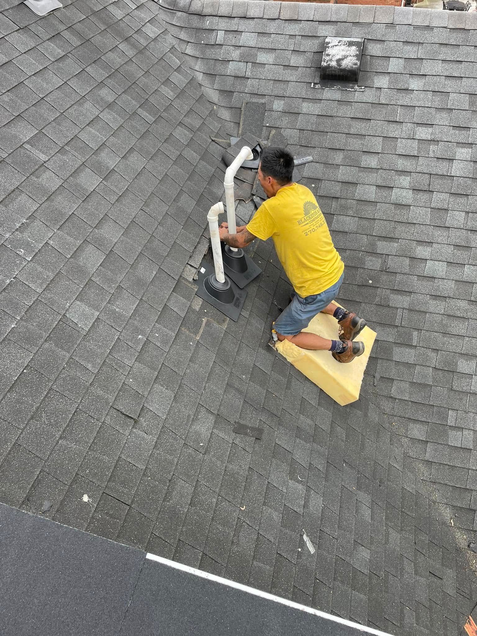 Worker in a yellow shirt kneeling on a roof, repairing a vent pipe on gray shingles.