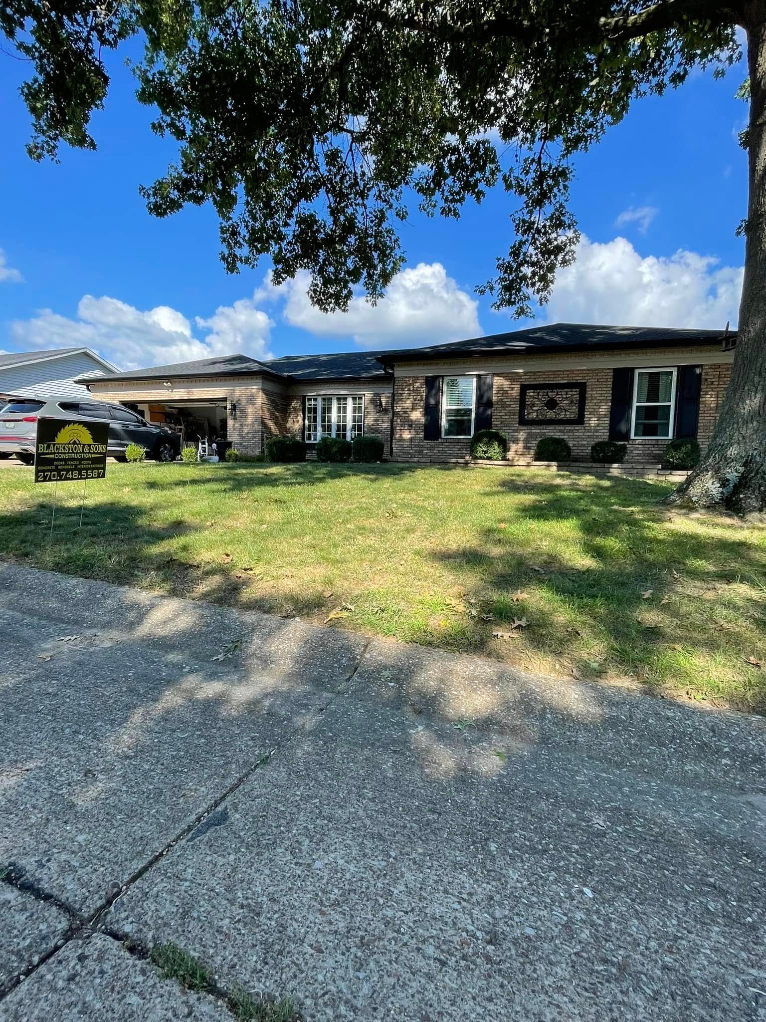 Single-story brick house with black shutters, gravel driveway, and a tree casting shade on the lawn.