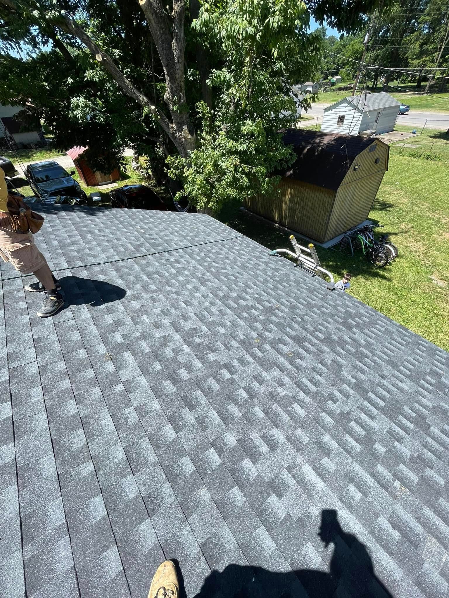 Close-up of a gray shingled roof with tree branches and a yard in the background