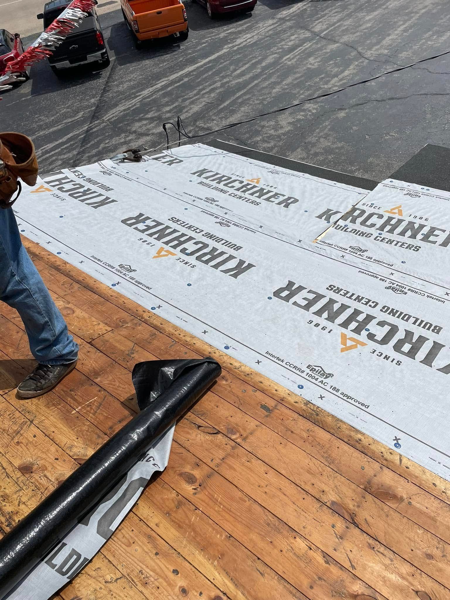 Worker installing white roofing underlayment on a sloped roof with orange shingles and tools nearby.