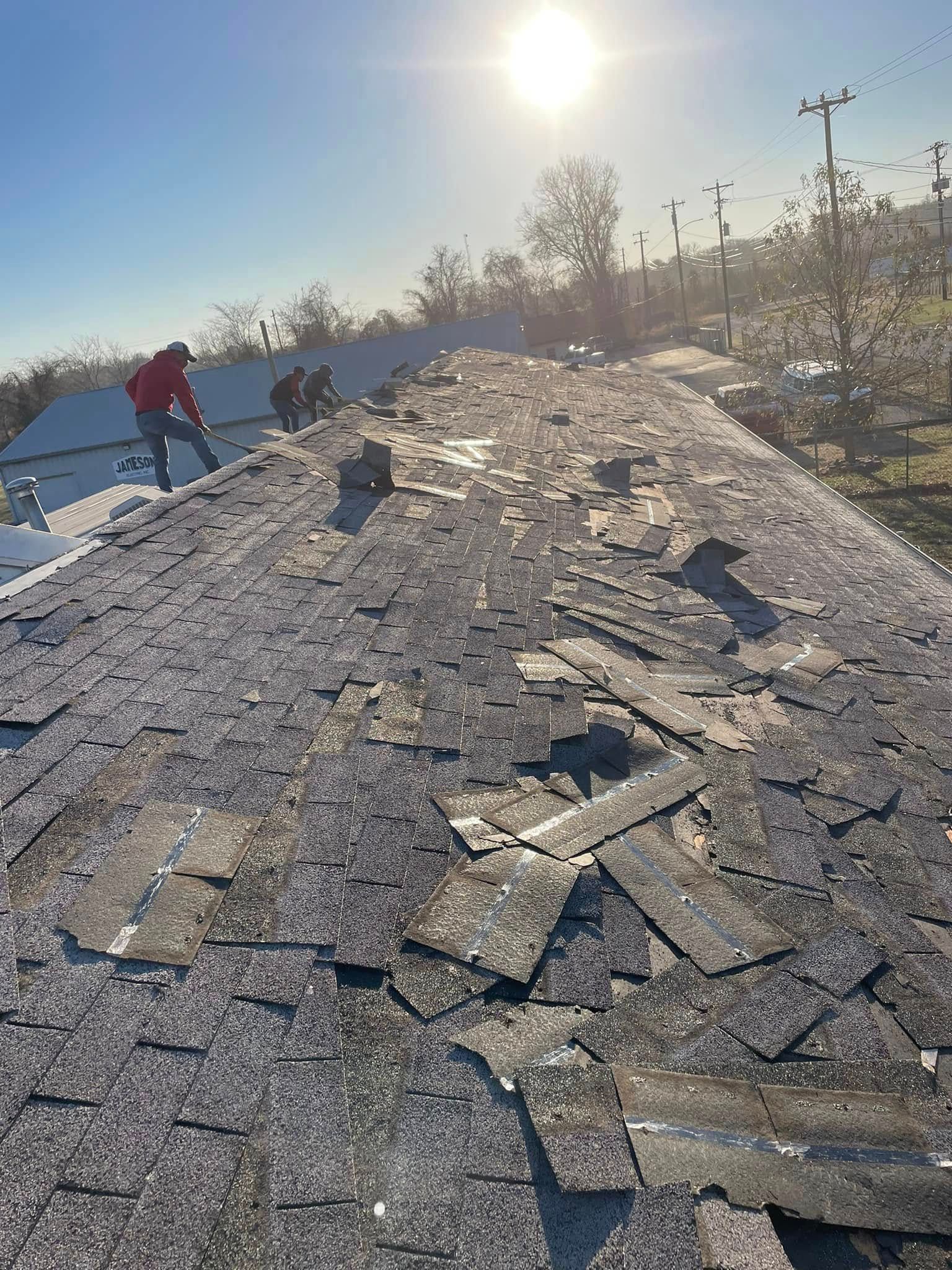 Workers on a shingled roof under bright sun, with solar panels and trees in the background.