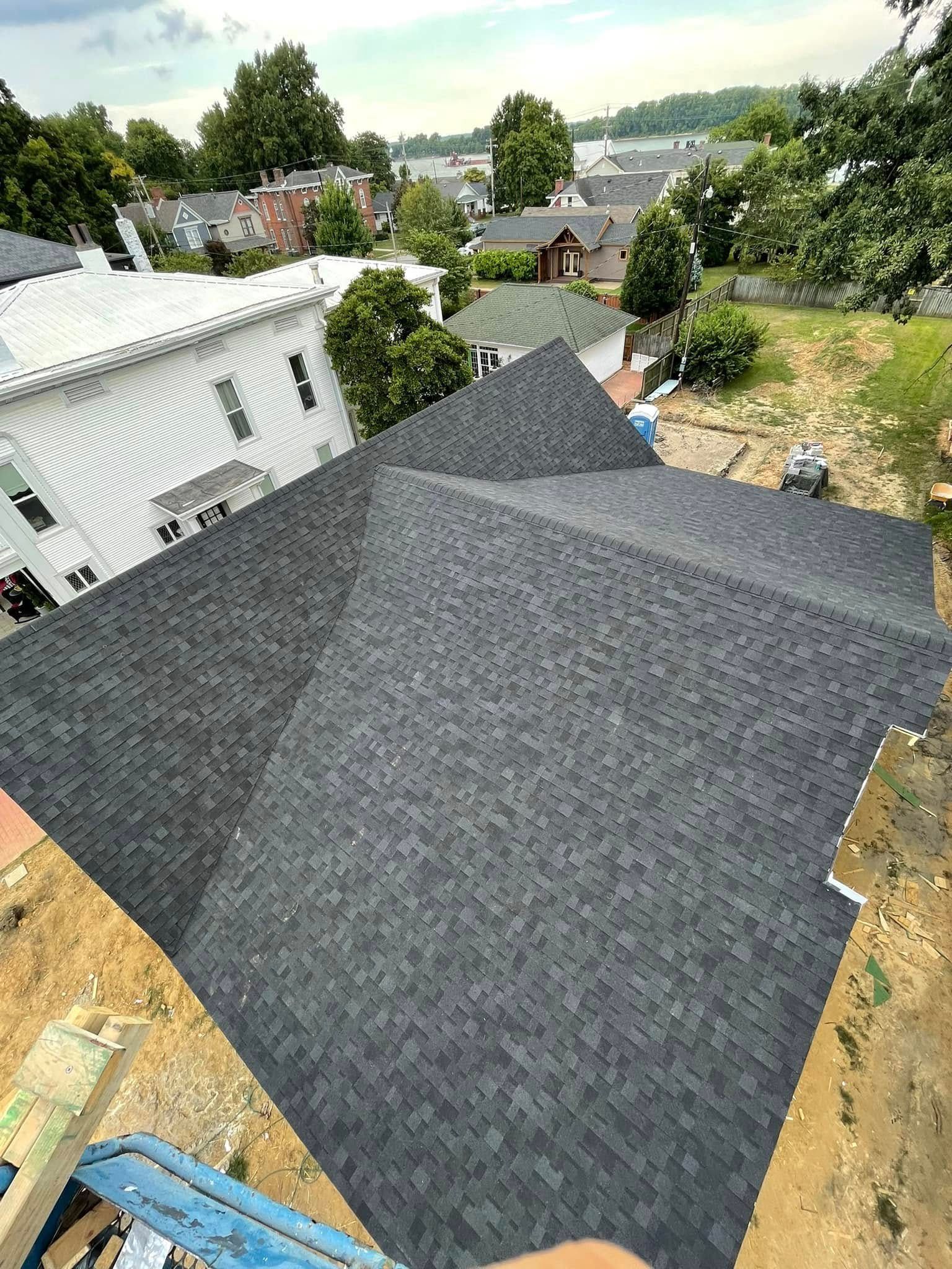 Dark gray roofing shingles on a steep roof under construction, with neighboring houses and trees in the background