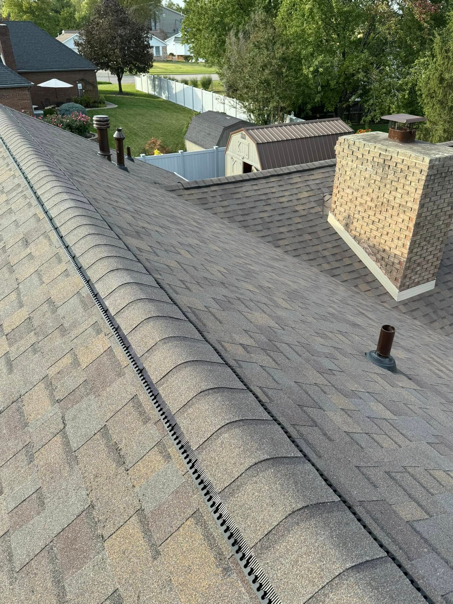 Rooftop covered in gray shingles with vents, chimneys, and trees in the background.