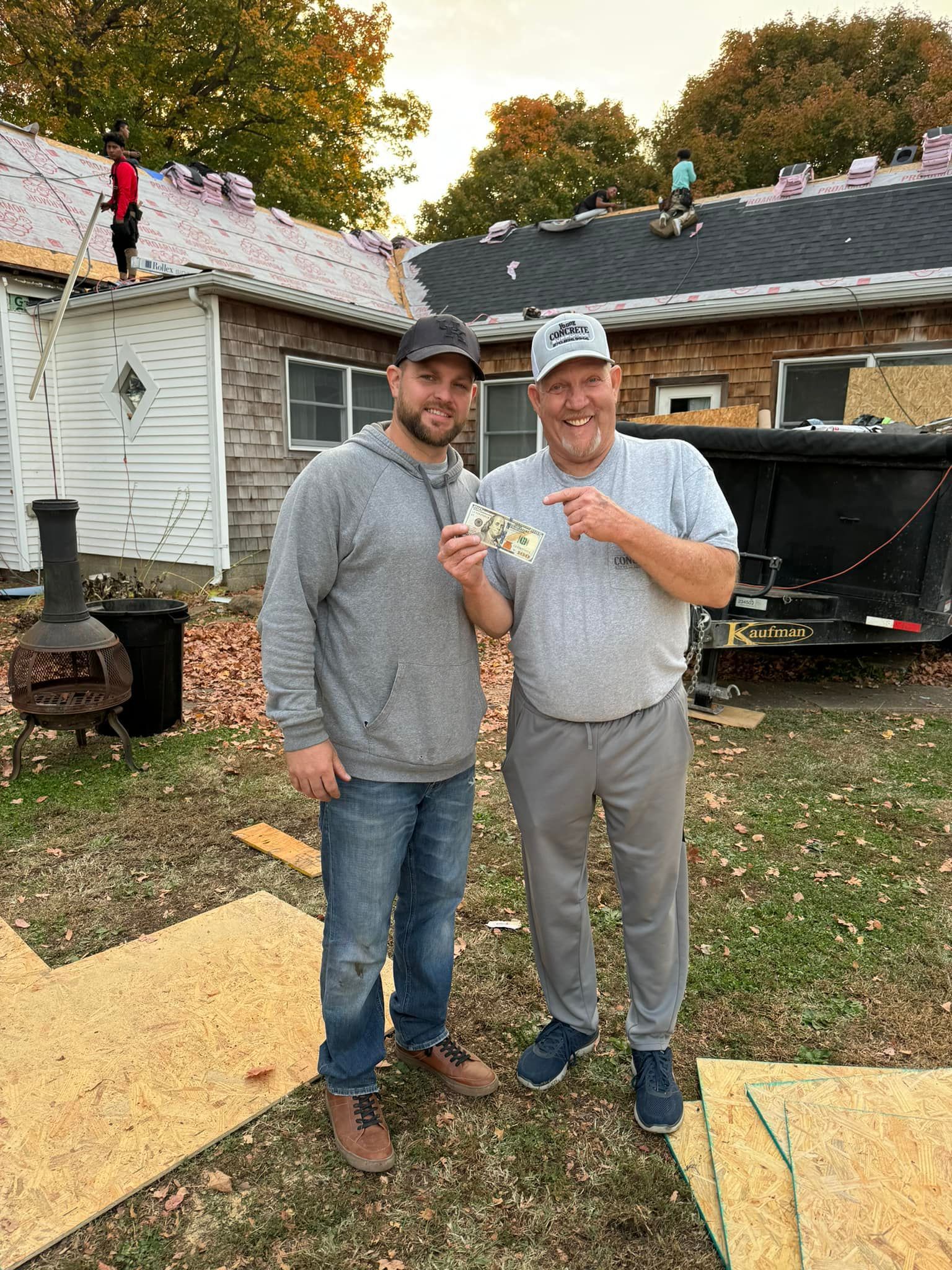Two men stand outdoors holding drinks in front of a house with a damaged roof and autumn leaves.