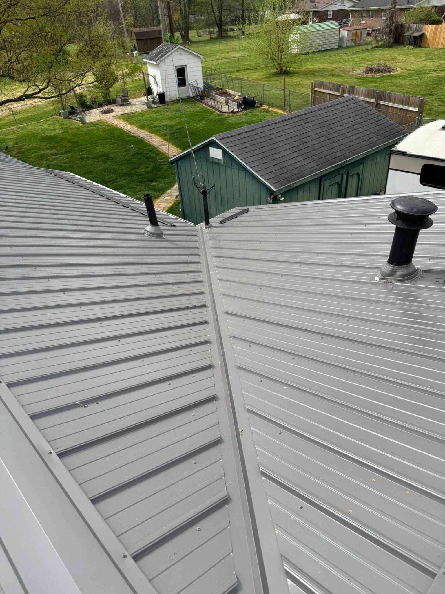 Rooftop view of gray metal roofs, a green shed, a small white outbuilding, and a grassy yard with trees.