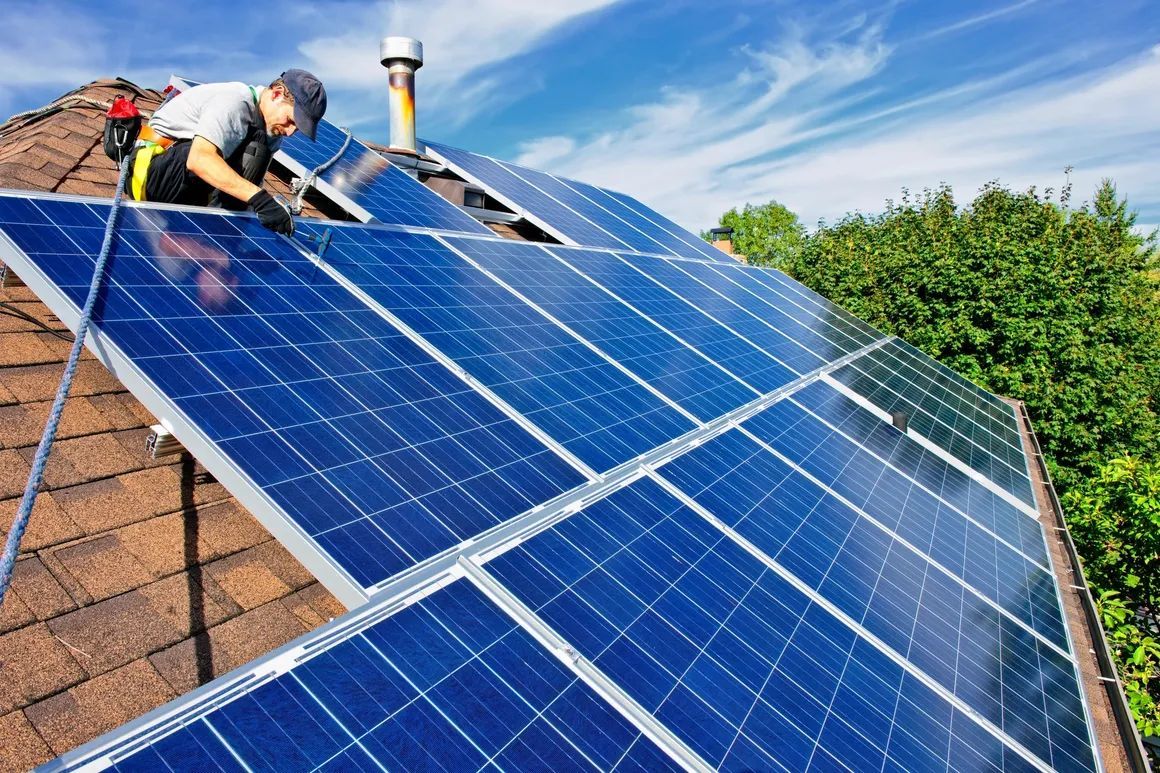 Worker installing blue solar panels on a rooftop under a bright sky